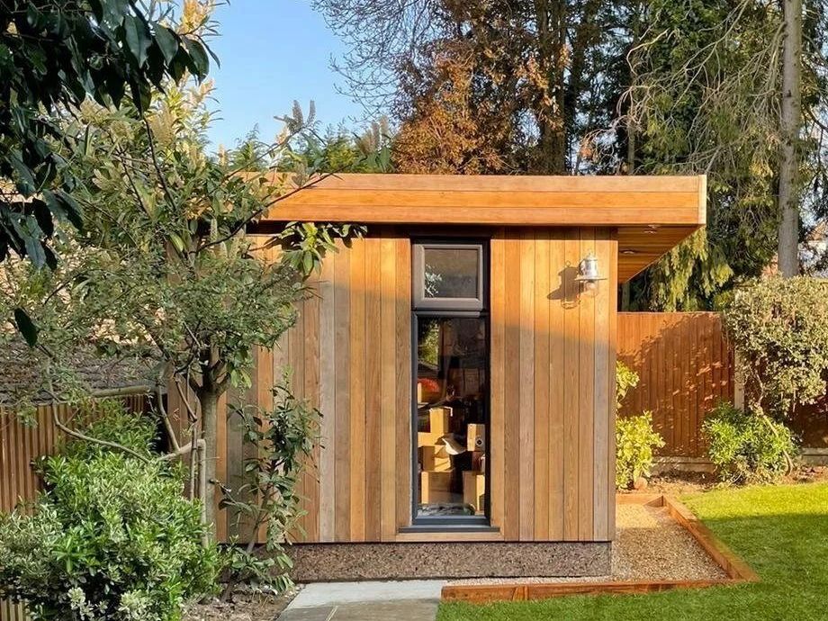 Same cedar garden office seen down a stone path, framed by shrubs and trees, lawn to the right