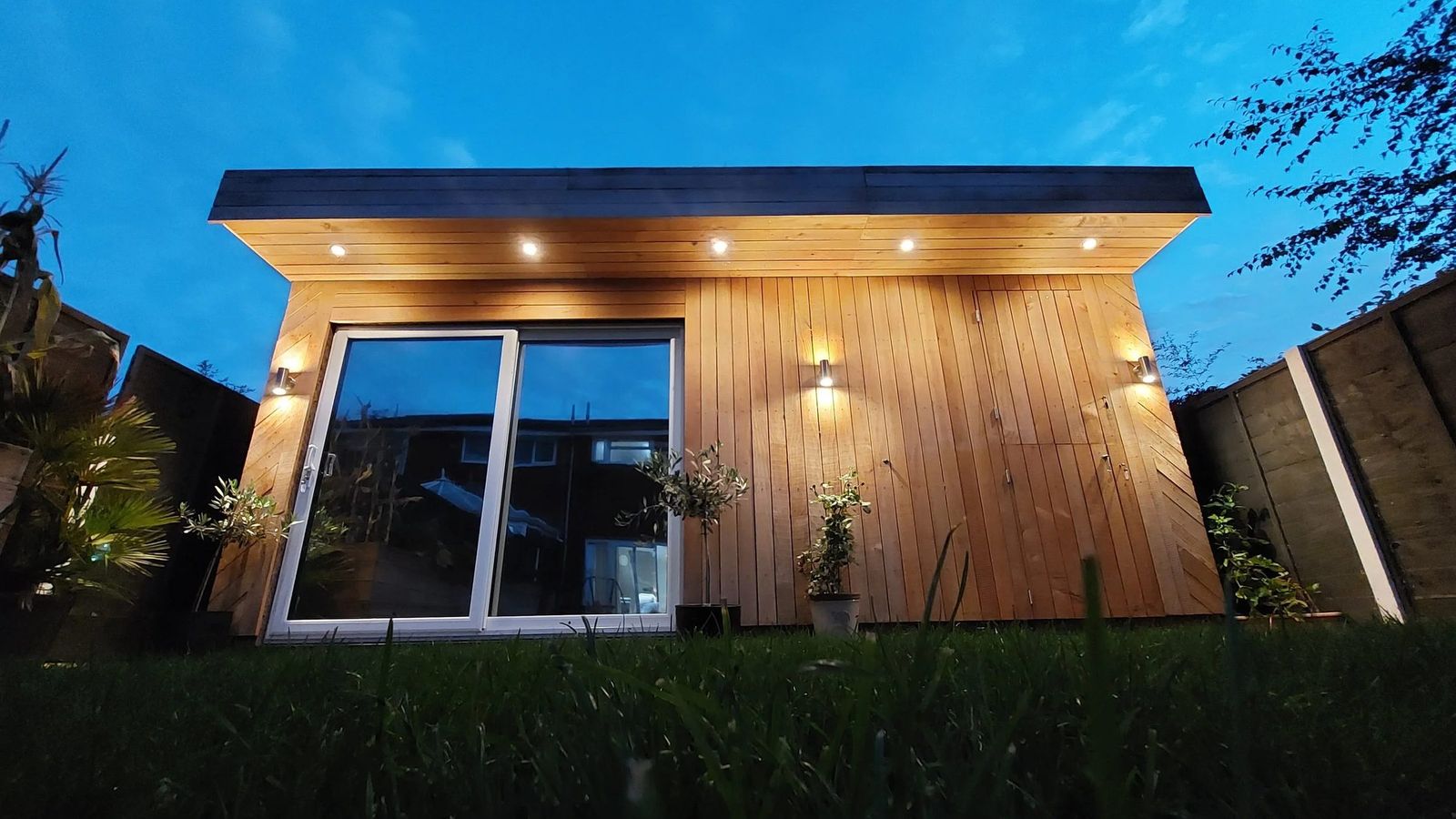 Cedar-clad flat-roof garden studio at dusk, low angle across long grass