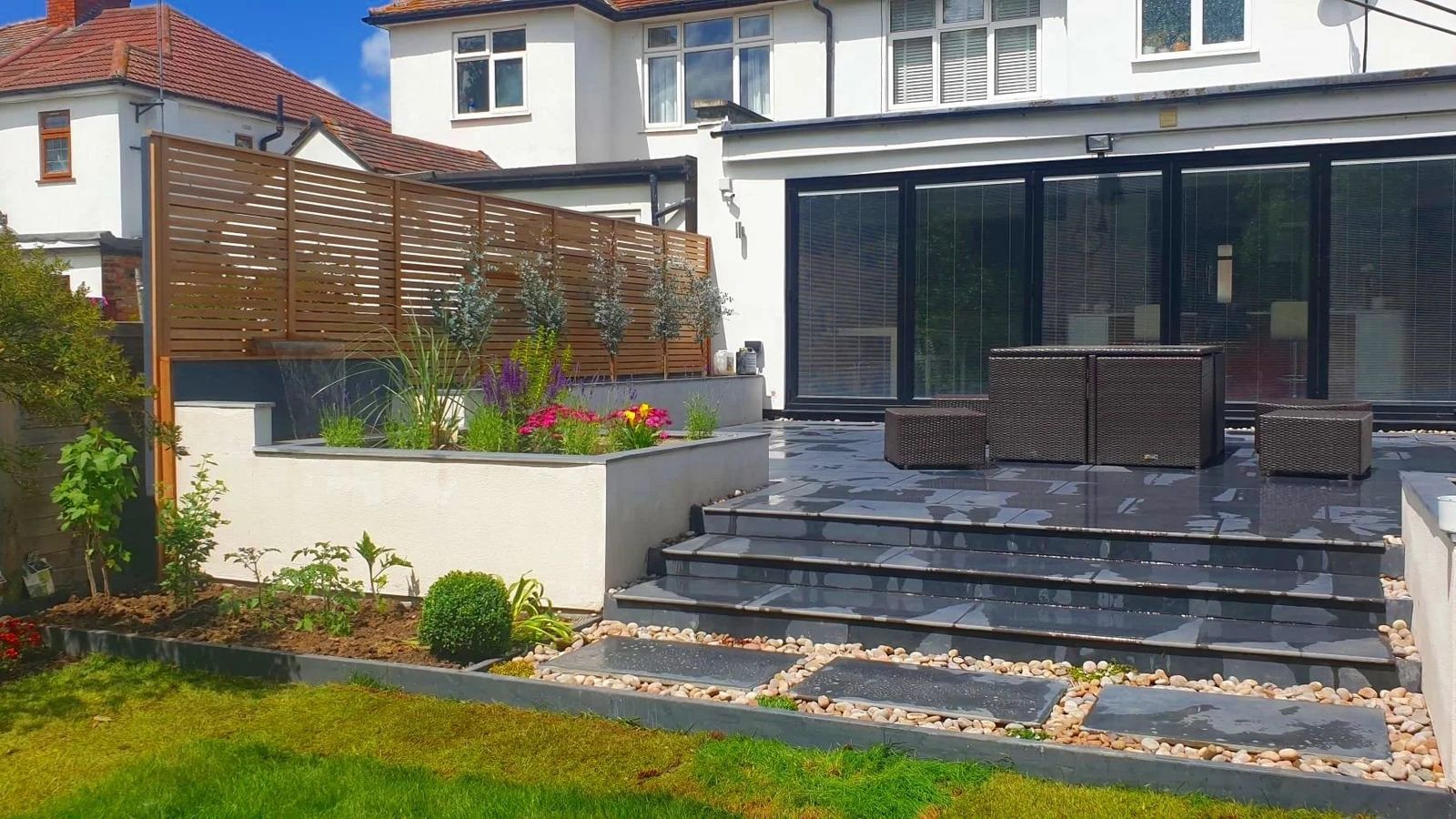 Rear of a white-rendered house with black bifold doors, dark slate steps and terrace with rattan dining set